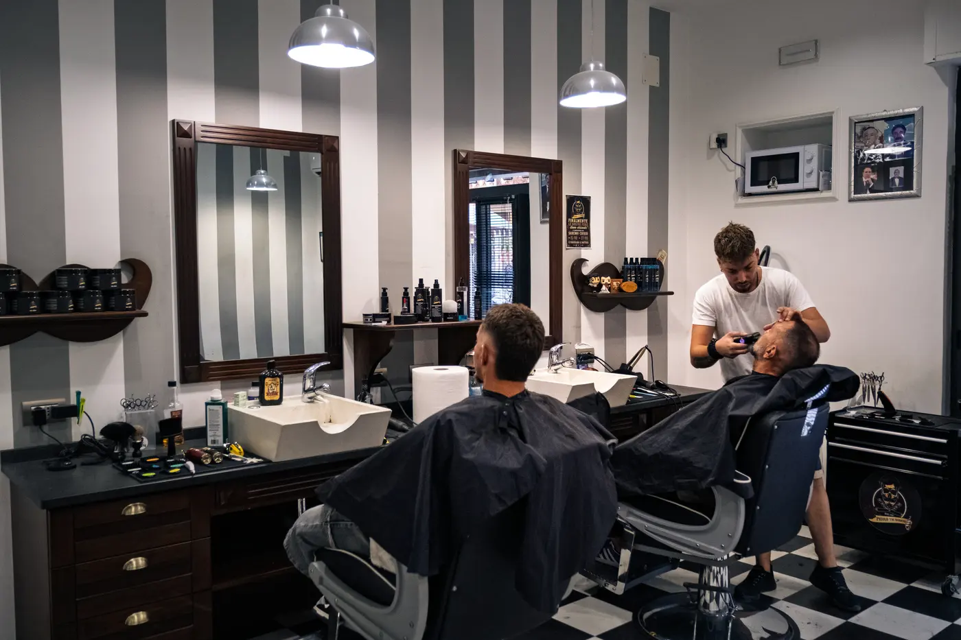 La sala di Priolo the Barbers: pavimento a scacchi bianchi e neri, due barbieri al lavoro insieme sulle poltrone.